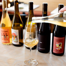 Load image into Gallery viewer, Wine bottles on a wooden table with one being poured into a glass.