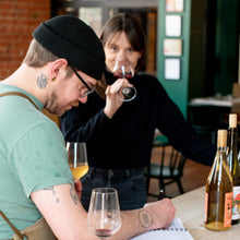 Load image into Gallery viewer, Two people tasting wine indoors with bottles and glasses on a table.